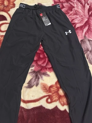 Svarta träningsbyxor från Under Armour - Svarta träningsbyxor från Under Armour med elastisk midja och dragsko. Byxorna har en vit logga på låret och är tillverkade i ett lätt och funktionellt material som passar perfekt för sport och träning.