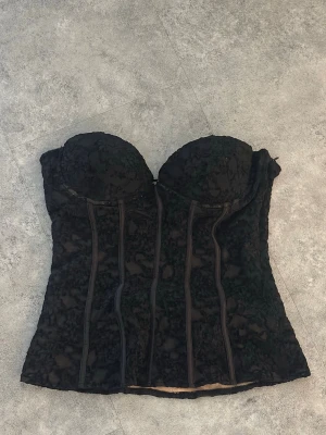 Svart spetskorsett  - Säljer en snygg svart korsett med vadderade kupor och transparent spetsdetaljer. Korsetten har markerade sömmar framtill för en formande effekt och nude-färgad insida. Axelbandslös design och dragkedja i sidan.
