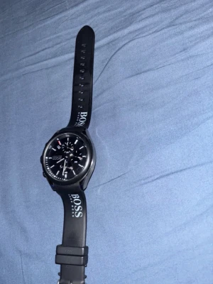 Svart Hugo Boss klocka med gummiband - Stilren svart klocka från Hugo Boss med svart urtavla och vita detaljer. Klockan har ett svart gummiband med tydlig BOSS-logga och flera funktioner på urtavlan. Perfekt accessoar för dig som gillar en sportig och modern look. BATTERIET ÄR DÖD, BEHÖVS ATT BYTA UT