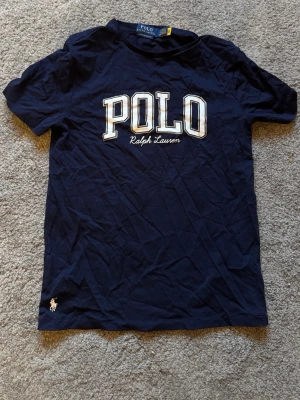 Marinblå POLO t-shirt från Ralph Lauren - Snygg marinblå t-shirt från Ralph Lauren med stort POLO-tryck och broderad logga vid nederkanten. Klassisk rund hals och korta ärmar. Tillverkad i mjuk bomull som känns skön mot huden. Perfekt för en clean och sportig look.