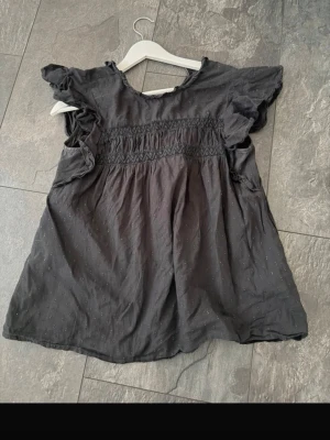 Svart blus med volangärmar - Söt svart blus med volangdetaljer vid ärmarna. blusen har en lätt struktur i tyget och är perfekt för dig som gillar en avslappnad men ändå feminin stil. Passar lika bra till sneakers som till boots.