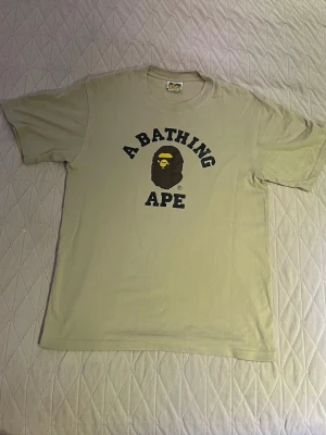 Beige Bape t-shirt - Säljer en beige t-shirt från A Bathing Ape med det klassiska aptrycket och texten 'A BATHING APE' i svart på bröstet. T-shirten har rund hals och korta ärmar. Perfekt för dig som gillar streetwear och vill sticka ut.