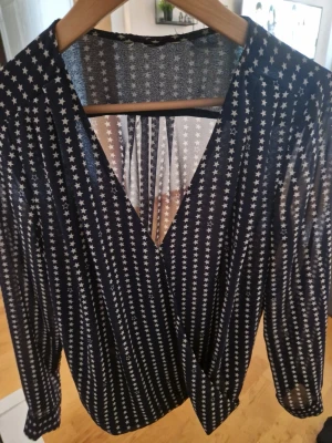 Blå blus med vita stjärnor - Cool blå blus med vita stjärnor och djup v-ringning. Blusen har långa ärmar och är gjord i ett tunt, transparent material som ger en snygg och luftig känsla. Perfekt för dig som gillar att sticka ut med mönster och detaljer.