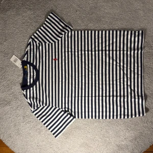 Randig t-shirt från Ralph Lauren - Snygg t-shirt med marinblå och vita ränder från Ralph Lauren. Klassisk passform med korta ärmar och rund halsringning med mörkblå kant. Liten röd broderad logga på bröstet. Perfekt för en stilren och avslappnad look.