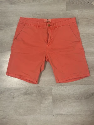 Korallfärgade shorts från A&F - Snygga korallfärgade shorts från Abercrombie & Fitch med klassisk passform. Shortsen har två sidofickor, två bakfickor och bälteshällor. Perfekta för varma dagar och ger en fräsch look till din outfit.