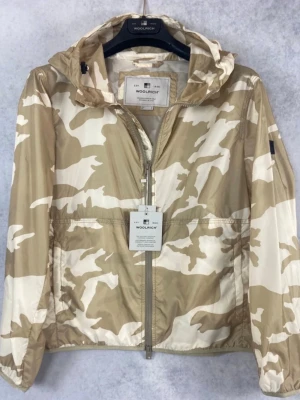 INTRESSEKOLL. Beige camo vindjacka från Woolrich.  - Iktressekoll på denna. Snygg vindjacka från Woolrich i beige och vitt camomönster. Jackan har huva, dragkedja framtill och två sidofickor. Tillverkad i lätt och tåligt material som passar perfekt för blåsiga dagar. Stilsäker look med avslappnad passform och diskret logga på ärmen.. 