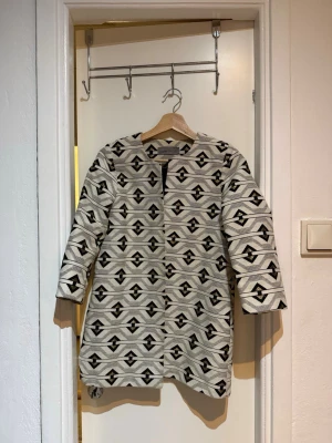 Mönstrad rock från Vero Moda - Snygg rock från Vero Moda i beige och svart med geometriskt mönster och inslag av guld. Rak passform, trekvartsärmar och öppen framtill utan synliga knappar. Tjockare tyg som ger en cool och modern look.