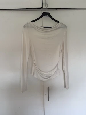 Vit långärmad topp med drapering - Stilren vit långärmad topp med lätt draperad halsringning och figurnära passform. Tillverkad i ett mjukt, stretchigt material som ger en elegant siluett. Perfekt att matcha med både jeans och kjol för en trendig look.