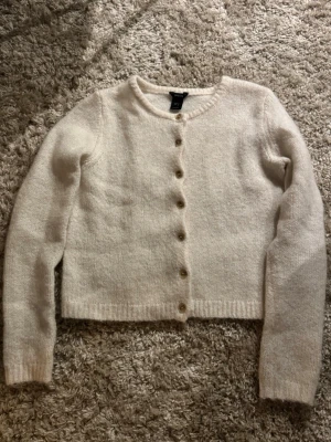 Vit kofta från Lindex  - Mysig beige kofta från Lindex  med rund halsringning och långa ärmar. Koftan har ribbade muddar och knäpps med åtta små knappar framtill. Perfekt att slänga över en topp för en enkel och stilren look.