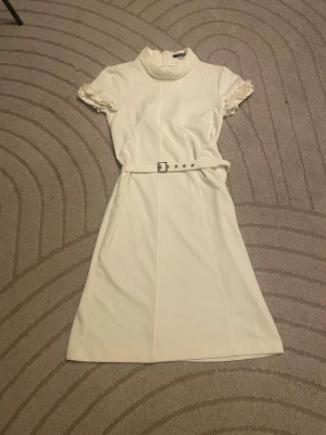 Vit kortklänning med bälte - Snygg vit kortklänning med hög krage och korta puffärmar med spetsdetaljer. Klänningen har ett matchande bälte i midjan och en enkel, stilren design som ger en klassisk look. Perfekt för dig som gillar en feminin och elegant stil.