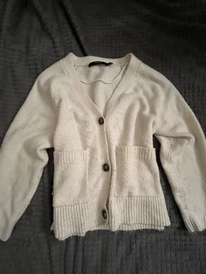 Vit kofta med knappar och fickor - Mysig vit kofta med v-ringning, stora knappar framtill och två rymliga fickor. Koftan har lång ärm och ribbade detaljer vid fickor, ärmslut och nederkant. 