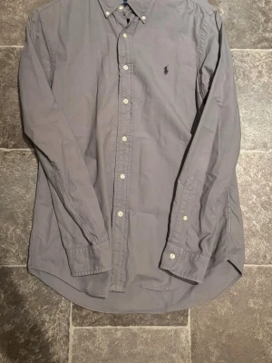 Grå skjorta från Ralph Lauren - Klassisk grå skjorta från Ralph Lauren med broderad logga på bröstet. Skjortan har lång ärm, knappar framtill och en tidlös design som passar till allt. Tillverkad i mjuk bomull för en bekväm känsla.