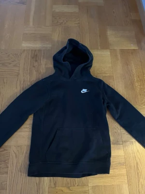 Svart hoodie från Nike - Svart hoodie från Nike med klassisk vit logga på bröstet. Hoodien har en stor huva och en rymlig magficka framtill. Tillverkad i mjukt material som känns skönt mot huden. Perfekt för en avslappnad och sportig stil.