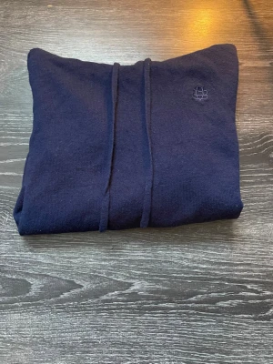 Mörkblå hoodie från Bvaldi  - Mörkblå hoodie från Bvaldi med broderad logga på bröstet. Tillverkad i mjukt stickat material, har huva med dragsnören och ribbade muddar vid ärmslut och nederkant. Perfekt för en avslappnad och stilren look.