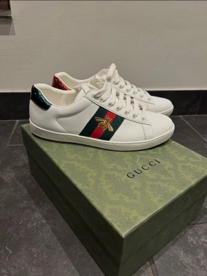 Gucci Ace vita sneakers med bi - Säljer ett par vita Gucci Ace sneakers i läder med klassiska gröna och röda ränder på sidan samt en broderad guldfärgad bi. Skorna har metallic-detaljer i rött och grönt på hälen och vita snören. Perfekta för dig som vill sticka ut med lyxig streetstyle.