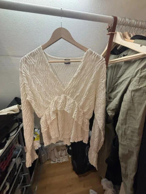 Vit omlottopp med volang - Säljer en vit omlottopp med lång ärm och volangdetalj framtill. Toppen har en v-ringad skärning och är gjord i ett strukturerat tyg som ger en unik look. Perfekt för dig som vill ha något extra till din outfit. Från Gina Tricot!