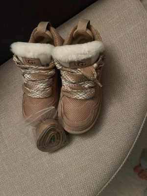 Beige sneakers från UGG  - Snygga beige sneakers från UGG med grova vita och beige skosnören och fluffig fuskpäls runt kanten. Ovandelen är i textil och mockaliknande material. Kommer med extra skosnören. Perfekta för dig som vill ha både stil och komfort.