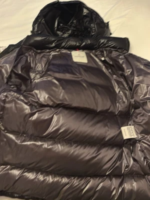 Svart dunjacka från Moncler - Snygg svart dunjacka från Moncler med glansig finish och klassisk puffer-design. Jackan har huva, dragkedja och innerficka samt ett unikt serietidningsmärke på insidan. Perfekt för kalla vinterdagar och riktigt varm tack vare dunfyllningen.
