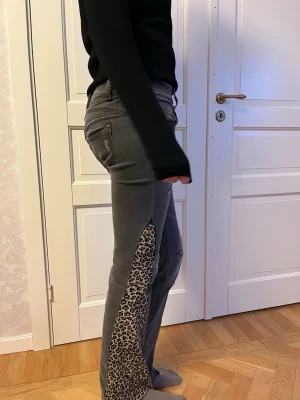 Lågmidjade jeans med leopardmönstrade slitsar - Unika grå, lågmidjade bootcut jeans  med leopardmönstrade slitsar längs sidorna. Jeansen har låg midja, fem fickor och slitna detaljer framtill. Perfekta för dig som vill spicea upp dina outfits