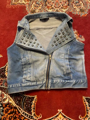Blå jeansväst med nitar - Säljer en blå jeansväst med coola silverfärgade nitar på kragen och dragkedja framtill. Västen har två fickor och en klassisk krage. Perfekt för att ge din outfit en edgy touch. Materialet är denim och färgen är ljusblå.