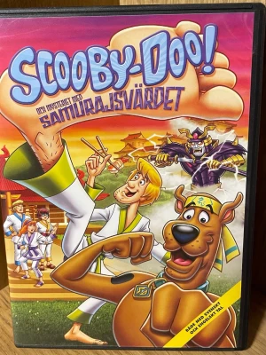 Scooby-Doo! och mysteriet med samurajsvärdet DVD - DVD-film med Scooby-Doo och gänget på äventyr i Japan där de löser mysteriet med ett magiskt samurajsvärd. Omslaget är färgglatt med tecknade karaktärer och japanska tempel i bakgrunden. Filmen har både svensk och engelsk tal.