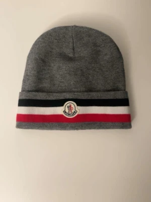 Grå Moncler mössa med logga - Snygg grå mössa från Moncler med klassisk logga framtill. Mössan har en uppvikt kant med ränder i svart, vitt och rött. Tillverkad i mjukt material som känns skönt mot huden. Perfekt accessoar för att lyfta din outfit.