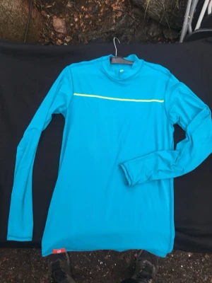 Blå långärmad träningströja Tenson - Säljer en blå långärmad träningströja från Tenson med en gul rand över bröstet. Tröjan har hög krage och är tillverkad i ett stretchigt funktionsmaterial som passar perfekt för löpning eller annan träning.