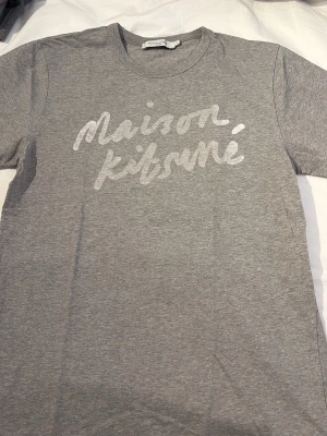 Grå Maison Kitsuné t-shirt - Snygg grå t-shirt från Maison Kitsuné med vit logotyp i handskriven stil på bröstet. Klassisk rund halsringning och korta ärmar. Tillverkad i mjuk bomull för en bekväm känsla och enkel att matcha med allt.Väldigt bra skick storlek M