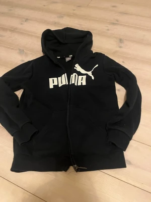 Svart hoodie med dragkedja från Puma - Svart hoodie från Puma med vit logga och text på bröstet. Tröjan har huva, dragkedja framtill och två fickor. Tillverkad i mjuk bomullsblandning, perfekt för en avslappnad stil.