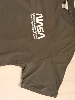  Grå NASA t-shirt med tryck - Cool grå t-shirt från NASA med stort tryck av NASA-loggan och text på bröstet samt en stor amerikansk flagga på ryggen. Klassisk passform och korta ärmar. Perfekt för dig som gillar rymden och vill ha en statement-tee.