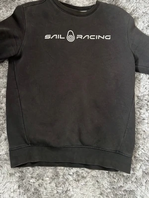 Sail Racing Sweatshirt  - Svart sweatshirt från Sail Racing med vit logga och text på bröstet. Klassisk rund halsringning och ribbade muddar vid ärmslut och nederkant. Perfekt för en avslappnad och stilren look.