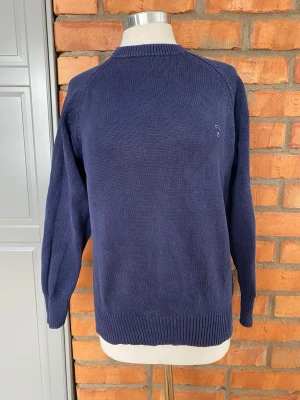 Mörkblå stickad tröja strl 42/44 - Klassisk mörkblå stickad tröja med rund halsringning och diskret logga broderad på bröstet. Tröjan har långa ärmar och ribbade muddar vid ärmslut och nederkant. Perfekt för lager på lager och enkel att matcha. 100% bomull.