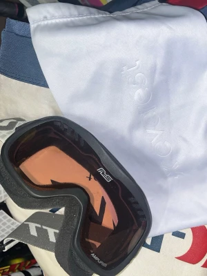 Svarta skidglasögon Everest Amplifier - Säljer ett par svarta skidglasögon från Everest med Amplifier-lins och RS-märkning. Glasögonen har rosa tonade linser och bekväm skumkant runt ramen. Perfekta för dig som vill ha bra sikt i backen. Förvaringspåse medföljer.