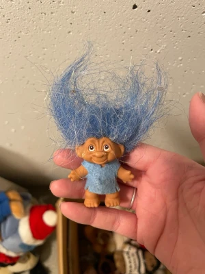Trollfigur med blått hår och tröja - Säljer en klassisk trollfigur med stort, rufsigt blått hår och en ljusblå tröja. Dockan är gjord av plast och har ett charmigt ansikte med stora ögon och utstående öron. Perfekt för samlare eller som rolig dekoration.