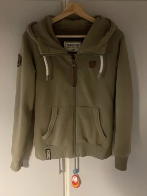 Beige hoodie från Naketano - Snygg beige hoodie från Naketano med dragkedja, stora fickor framtill och ribbade muddar. Huva med vita, tjocka dragsnören och detaljer i brunt konstläder. Tillverkad i mjukt sweatshirtmaterial, perfekt för chill dagar.