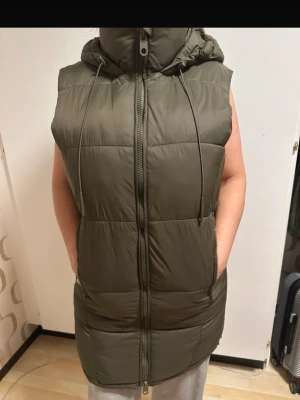 Olivgrön lång dunväst med huva - Snygg olivgrön dunväst med dragkedja framtill och stor huva. Västen är quiltad, har två fickor med dragkedja och dragsko i midjan för justerbar passform. Perfekt för lager-på-lager och ger en cool streetstyle-vibe.
