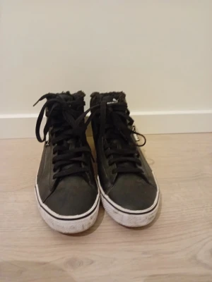 Svarta höga sneakers från Puma - Säljer ett par svarta höga sneakers från Puma med vit sula och svart pälskant runt öppningen. Skorna har klassisk snörning och Puma-logga på hälen. Materialet är en mix av syntet och textil, perfekta för dig som gillar streetstyle.