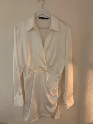 Vit omlottklänning från Zara - Stilren vit kortklänning från Zara med omlottdesign och djup v-ringning. Klänningen har långa ärmar, markerad midja och klassisk skjortkrage. Materialet har en lätt glansig finish.