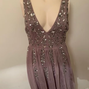 Lila långklänning med paljetter - Elegant lila långklänning med djup v-ringning och tunna axelband. Klänningen har glittriga paljettdekorationer på överdelen och ett flytande, skirt tyg i kjolen. Perfekt för speciella tillfällen där du vill sticka ut.