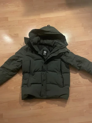  Canada Goose wyndham black label - Säljer en grå dunjacka från Canada Goose med huva och dragkedja samt knappar framtill. Jackan har två bröstfickor och två sidofickor, perfekt för kalla vinterdagar. Klassisk, stilren design och riktigt varm tack vare dunfyllningen.
