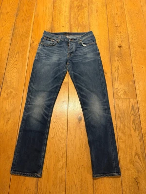 Nudie Jeans Grim Tim Cold Crisp - Säljer ett par klassiska blå jeans från Nudie Jeans med raka ben och snygga slitningar på framsidan. Jeansen har fem fickor, kontrastsömmar och detaljerade bakfickor med Nudie-logga. Tillverkade i bomull för en skön och hållbar känsla.