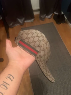 Äkta Gucci keps begie  - Säljer en beige keps från Gucci med det klassiska GG-monogrammet över hela kepsen. På sidan finns en ikonisk grön och röd rand. Kepsen har böjd skärm och justerbar rem bak. Materialet är canvas med exklusiv känsla.