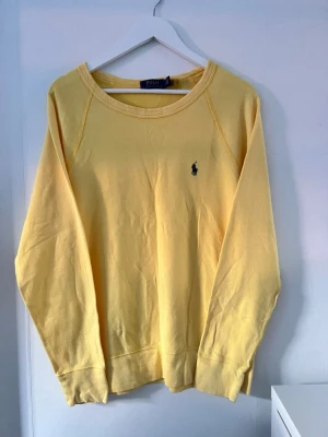 Gul sweatshirt från Polo Ralph Lauren - Gul sweatshirt från Polo Ralph Lauren med klassisk rund halsringning och broderad logga på bröstet. Tröjan har långa ärmar och är tillverkad i mjuk bomull, perfekt för en avslappnad och stilren look.