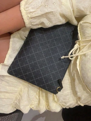 Svart kuvertväska med rutmönster - Stilren svart kuvertväska med diskret rutmönster och guldfärgad ringdetalj i hörnet. Väskan har en enkel, rektangulär form och stängs med snörning. Perfekt accessoar för att lyfta din outfit.