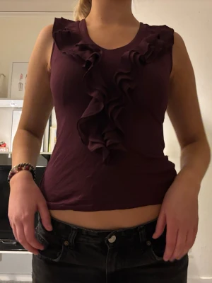 Mörkröd topp med volang - Snygg mörkröd topp med v-ringning och dekorativa volanger framtill. Perfekt för dig som vill ha en feminin och trendig look. Stretchigt material som sitter bekvämt och framhäver figuren.