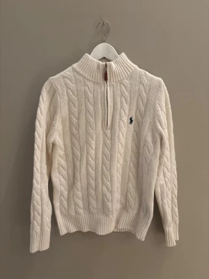 Vit kabelstickad tröja Polo Ralph Lauren - Kabelstickad vit tröja från Polo Ralph Lauren med halv dragkedja och hög krage. Tröjan har ribbade muddar och nederkant samt den klassiska broderade loggan på bröstet. Perfekt för dig som gillar en clean och tidlös look.