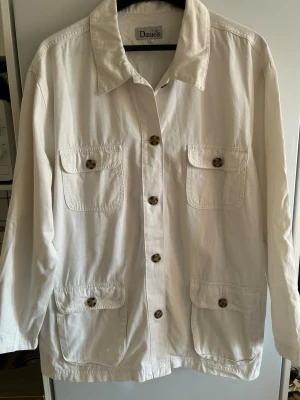 Vit overshirt från Daniels - Stilren vit overshirt från Daniels i bomull med fyra stora fickor framtill och klassisk krage. Knappar i kontrasterande färg ger en snygg detalj. Perfekt lager på lager-plagg med avslappnad passform och lång ärm.