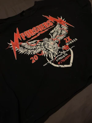 Svart croppad t-shirt med tryck - Cool svart croppad t-shirt med stort tryck av en örn och texten 'Pursuers' i rött och vitt framtill. T-shirten har korta ärmar och en edgy rockig vibe, perfekt för dig som gillar statement-plagg.