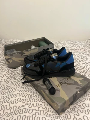 Valentino Garavani blå camo sneakers - Snygga sneakers från Valentino Garavani i svart och blått med camo-mönster. Skorna har svarta snören, robust svart sula och detaljer i läder och textil. Kommer med originalkartong i camo-design och extra skosnören.