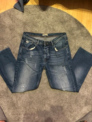 Blå jeans från Blend med slitningar - Säljer ett par blå jeans från Blend med snygga slitningar och tvättade detaljer. Klassisk femficksmodell med raka ben och normal midja. Jeansen har vita sömmar vid fickorna och ett läderpatch med logga bak i midjan. Perfekta för en avslappnad och trendig look.